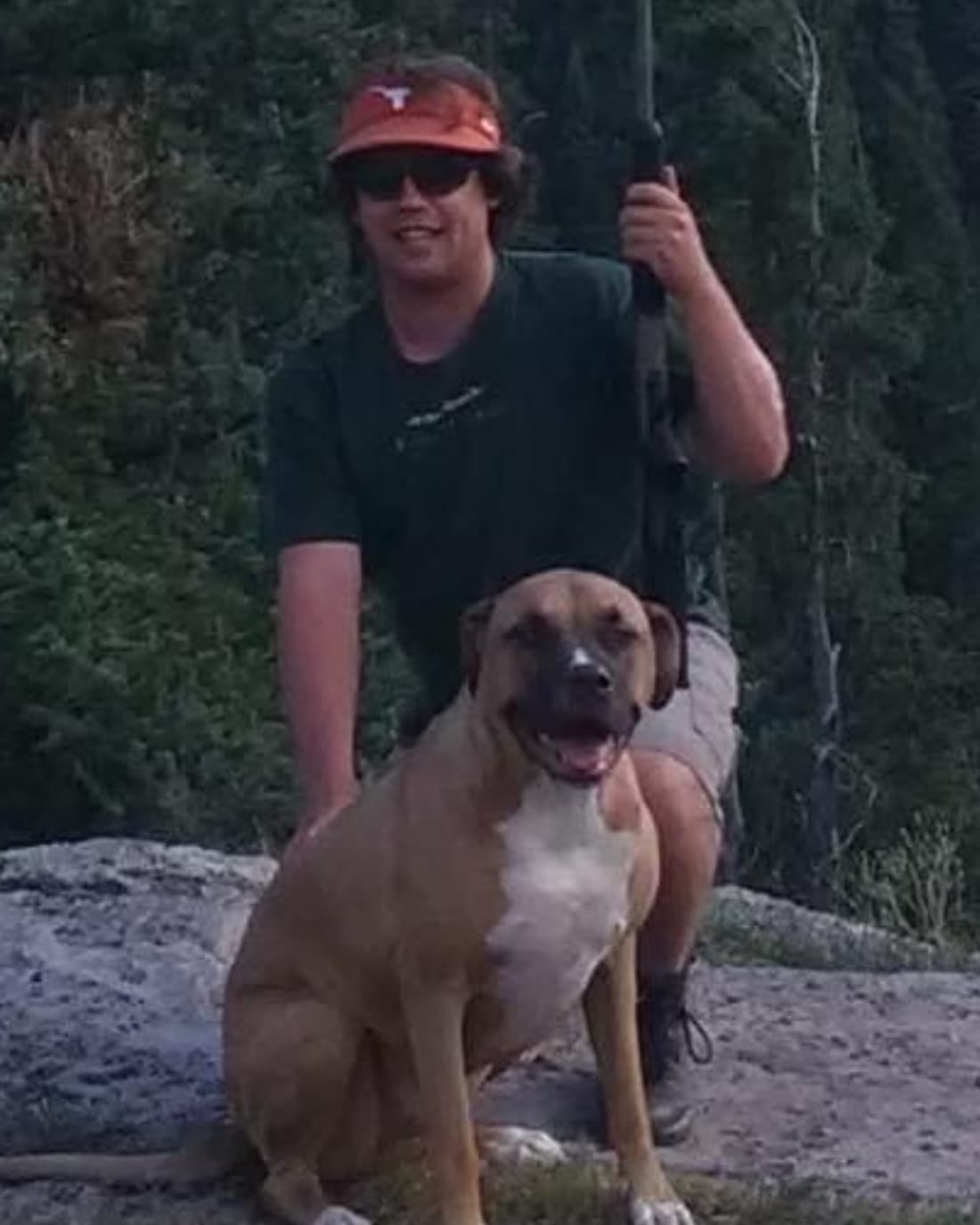 An angler in an orange visor kneeling on a granite slab with a fly rod, with Orbit, a tan dog, sitting in front, above Hermosa Creek