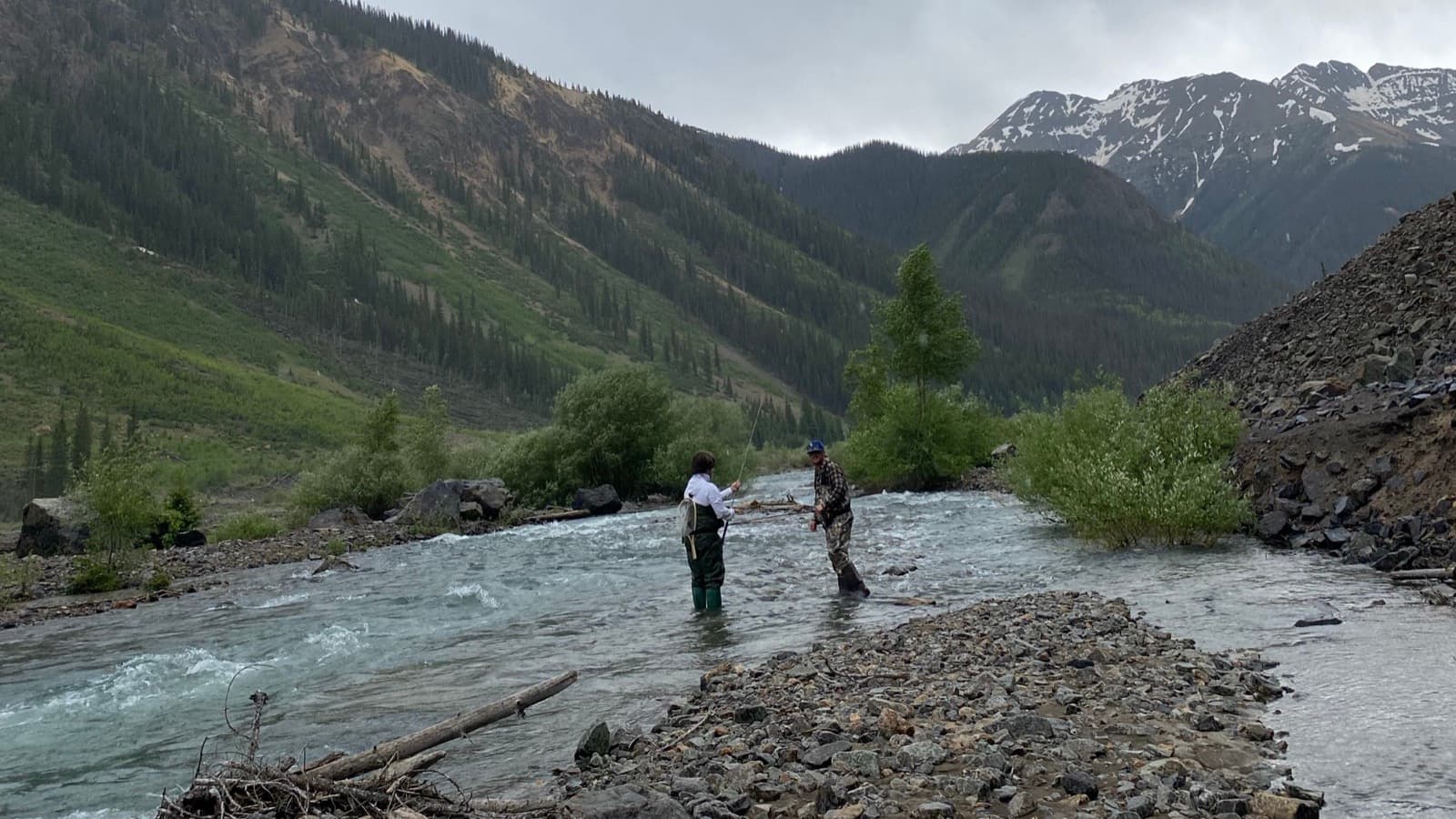 Two anglers wading the upper Animas River above Silverton, below snow capped peaks under a grey sky